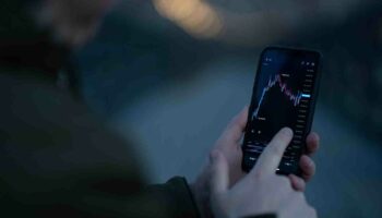 Businessman monitoring stock market data while trading online in mobile app Rear view of trader monitoring stock market data in mobile app for online trading and investing on smartphone while standing outdoors, selective focus on hand touching screen with forex graph chart